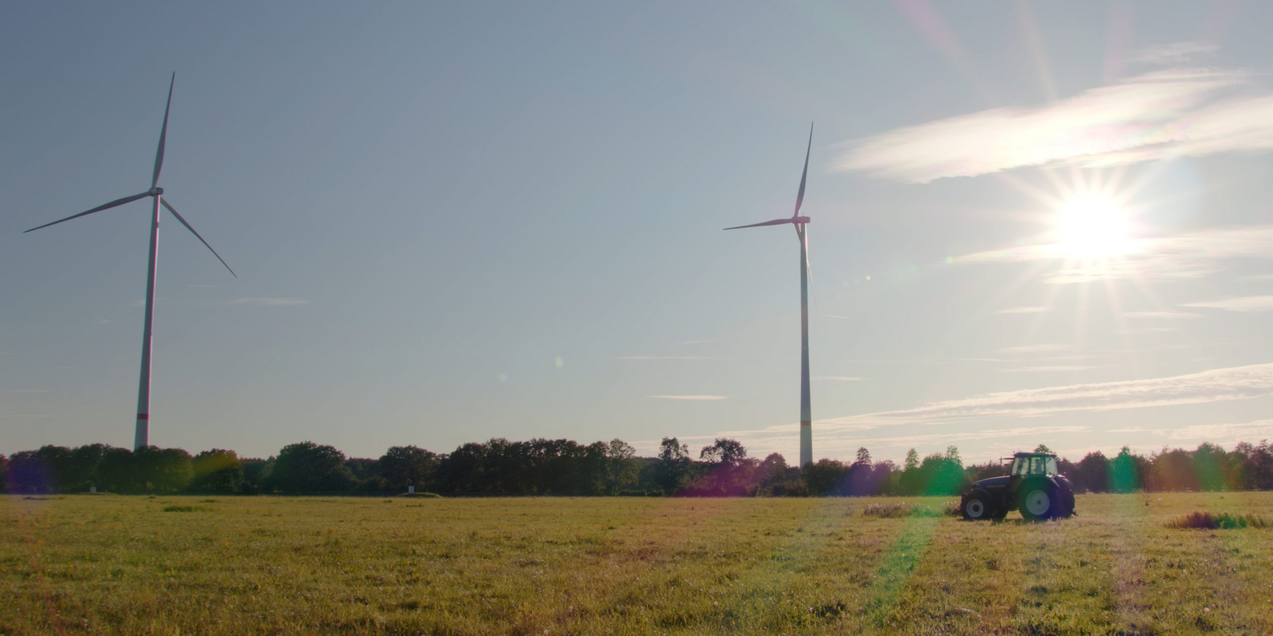 Als Symbolbild für das Projekt Windwende-Videos sieht man ein Foto zweier Windräder in Coesfeld bei Sonnenschein auf einem Feld mit einem Traktor davor.