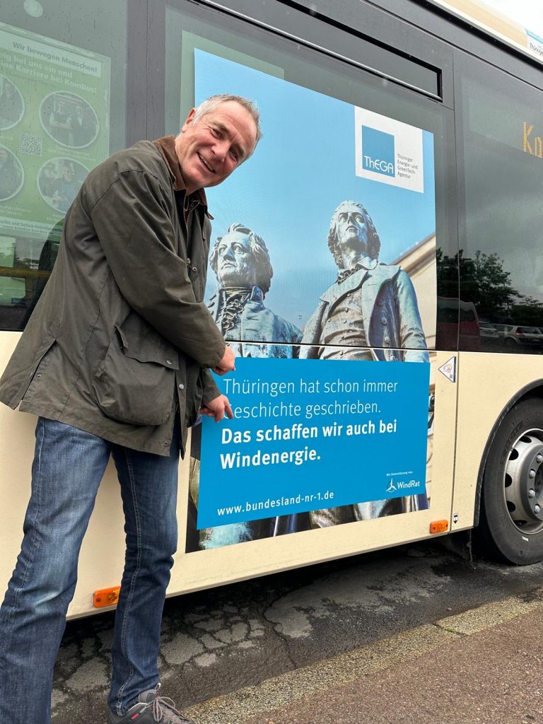 WindRat-Projektmanager Peter Keller steht vor einem Bus und deutet auf das Plakat, das auf den Bus geklebt ist: Das Kampagnen-Plakat mit der Aufschrift "Thüringen hat immer schon Geschichte geschrieben: Das schaffen wir auch bei Windenergie!". Weiterhin ist darauf die Statue von Schiller und Goethe und das Logo der Thüringer Landesenergieagentur und des WindRats zu sehen.