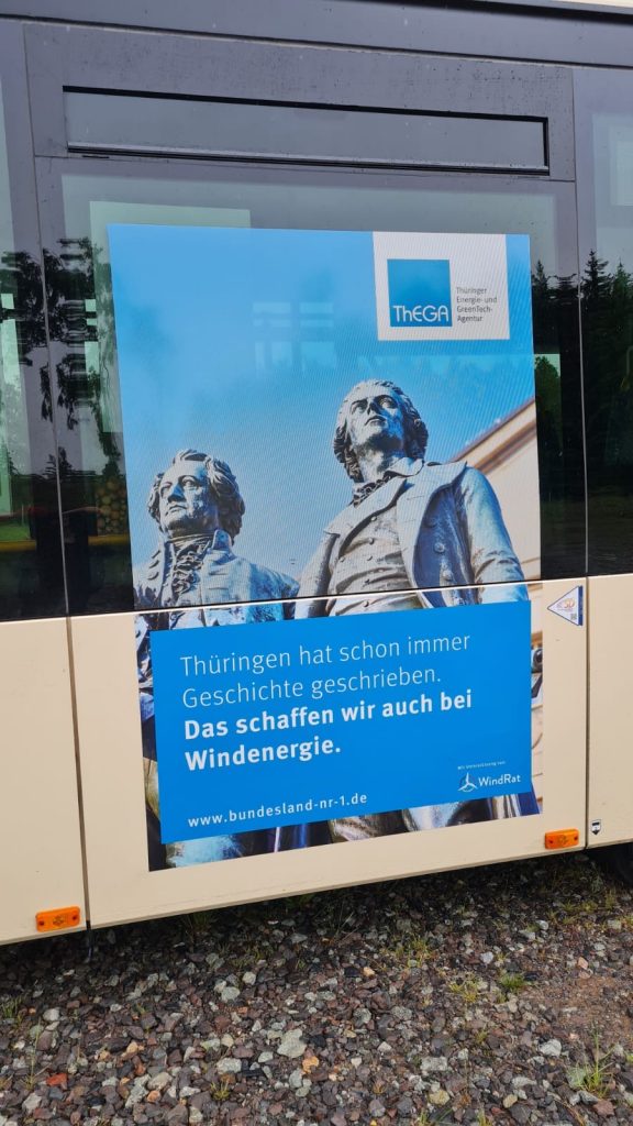 Man sieht ein Kampagnen-Plakat, das auf einem Bus aufgeklebt ist. Es trägt die Aufschrift "Thüringen hat immer schon Geschichte geschrieben: Das schaffen wir auch bei Windenergie!". Weiterhin ist darauf die Statue von Schiller und Goethe und das Logo der Thüringer Landesenergieagentur und des WindRats zu sehen.