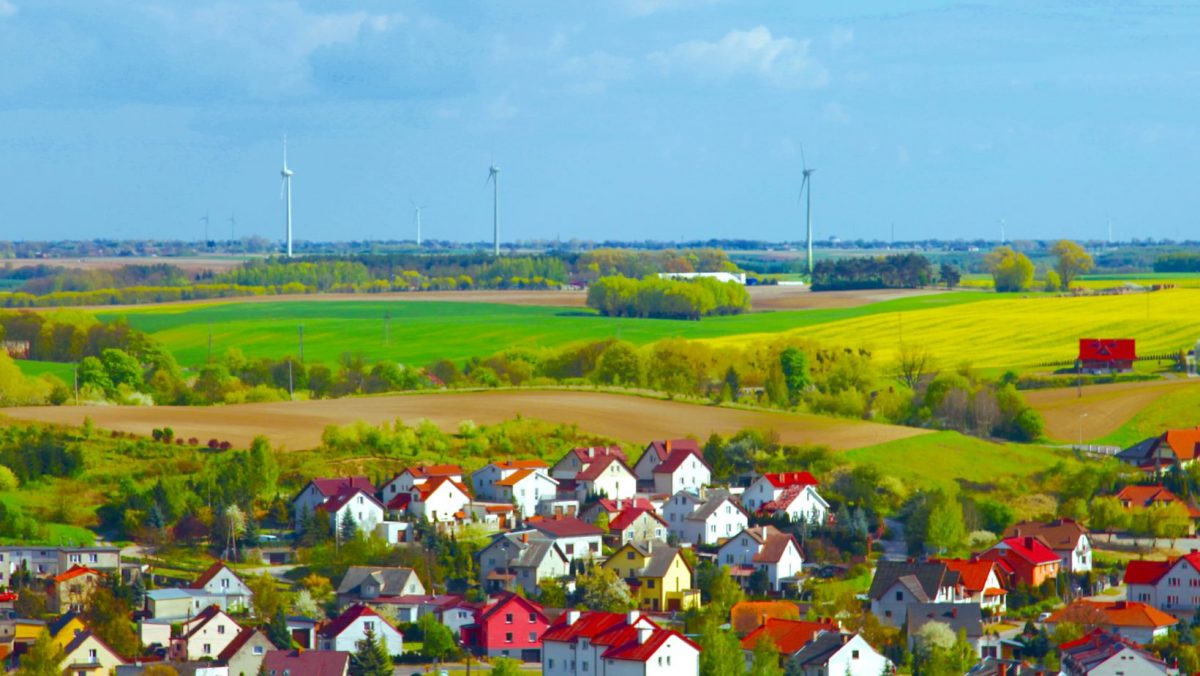 Man sieht eine kleine Stadt im Vordergrund vor weiten Feldern mit blauem Himmel und einigen Windenergieanlagen im Hintergrund.