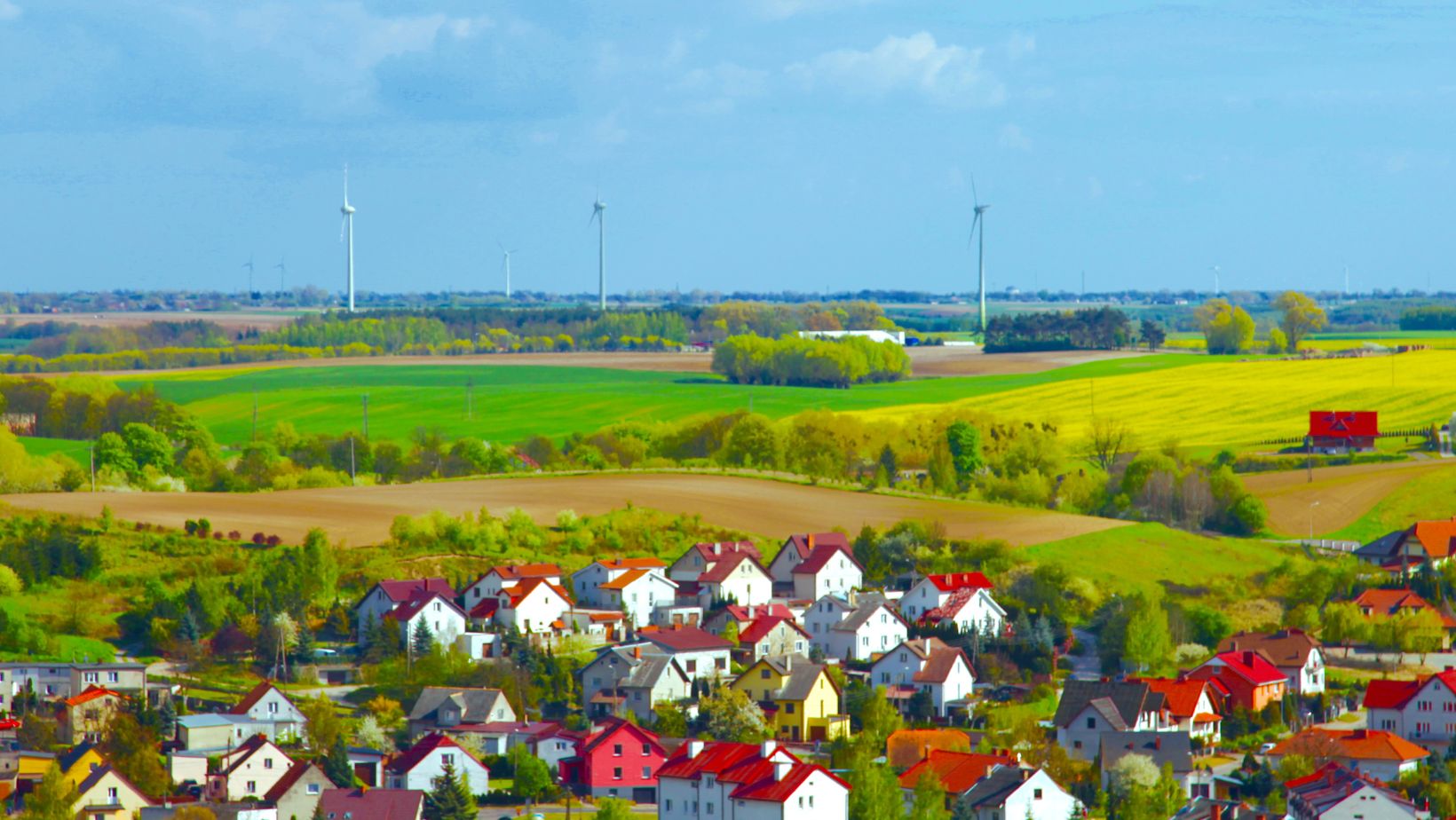 Man sieht eine kleine Stadt im Vordergrund vor weiten Feldern mit blauem Himmel und einigen Windenergieanlagen im Hintergrund.
