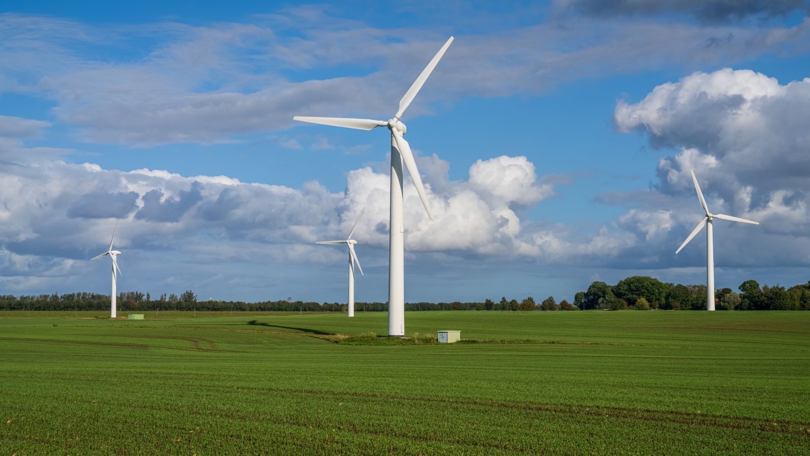 Man sieht vier Windräder auf grüner Wiese vor blauem Himmel mit Schäfchenwolken als Symbolbild für Windenergie als Wirtschaftsförderung