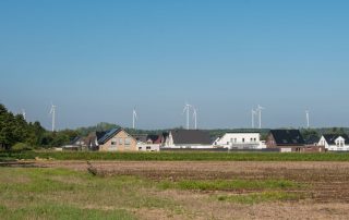 Acker mit kleiner Siedlung vor blauem Himmel mit einigen Windrädern im Hintergrund als Symbol für Umwelt- und Anwohnerschutz bei Windparkplanung.