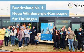 Eine Gruppe von ca. 20 Leuten steht bei trübem Wetter auf einer Wiese vor einem Bus. Im Hintergrund steht ein Windrad. Der Bus trägt das Kampagnen-Plakat mit der Aufschrift "Thüringen hat immer schon Geschichte geschrieben: Das schaffen wir auch bei Windenergie!". Weiterhin ist darauf die Statue von Schiller und Goethe und das Logo der Thüringer Landesenergieagentur und des WindRats zu sehen.
