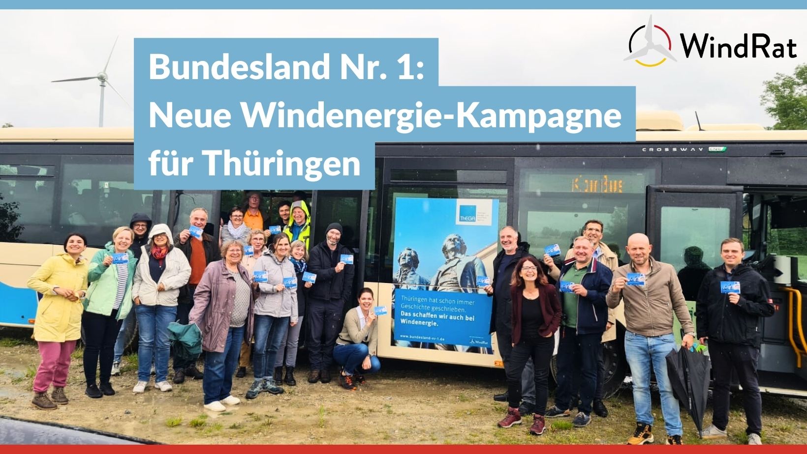 Eine Gruppe von ca. 20 Leuten steht bei trübem Wetter auf einer Wiese vor einem Bus. Im Hintergrund steht ein Windrad. Der Bus trägt das Kampagnen-Plakat mit der Aufschrift "Thüringen hat immer schon Geschichte geschrieben: Das schaffen wir auch bei Windenergie!". Weiterhin ist darauf die Statue von Schiller und Goethe und das Logo der Thüringer Landesenergieagentur und des WindRats zu sehen.