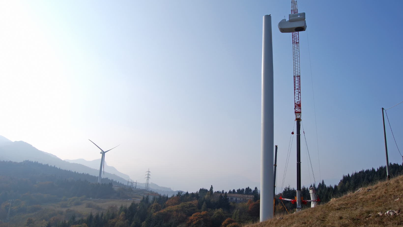 Eine Windenergieanlage im Bau mit Kran in der Natur, vor Bergen mit herbstlicher Bewaldung.