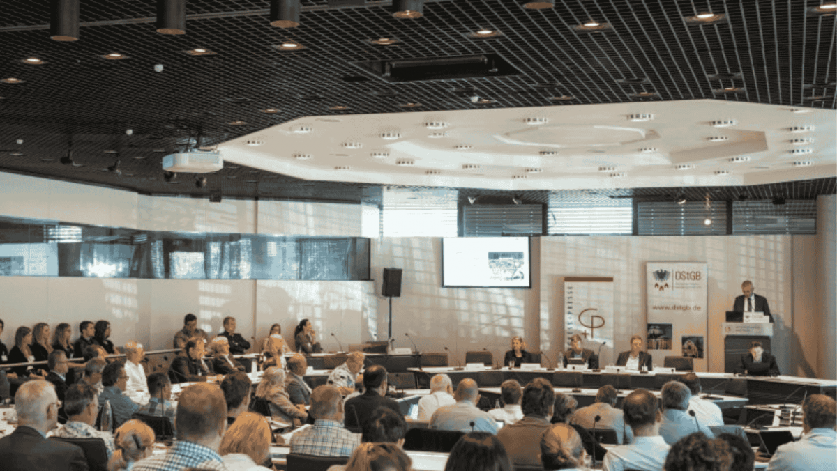 Man sieht einen Saal mit Podium, an dem ein älterer Herr einen Vortrag hält, und viele Menschen im Publikum. Es zeigt die 17. Fachkonferenz „Kommunen aktiv für den Klimaschutz“ am 14. Mai 2025 in Bonn.